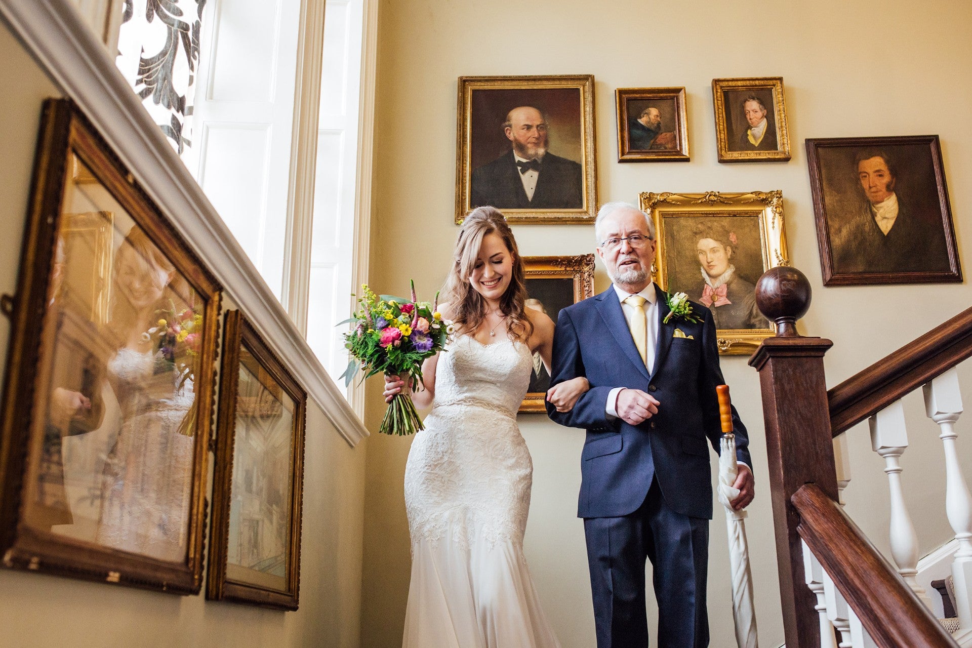 Bride carrying her floral bouquet linking arms with her dad as they walk down a wooden staircase at Ochardleigh House.