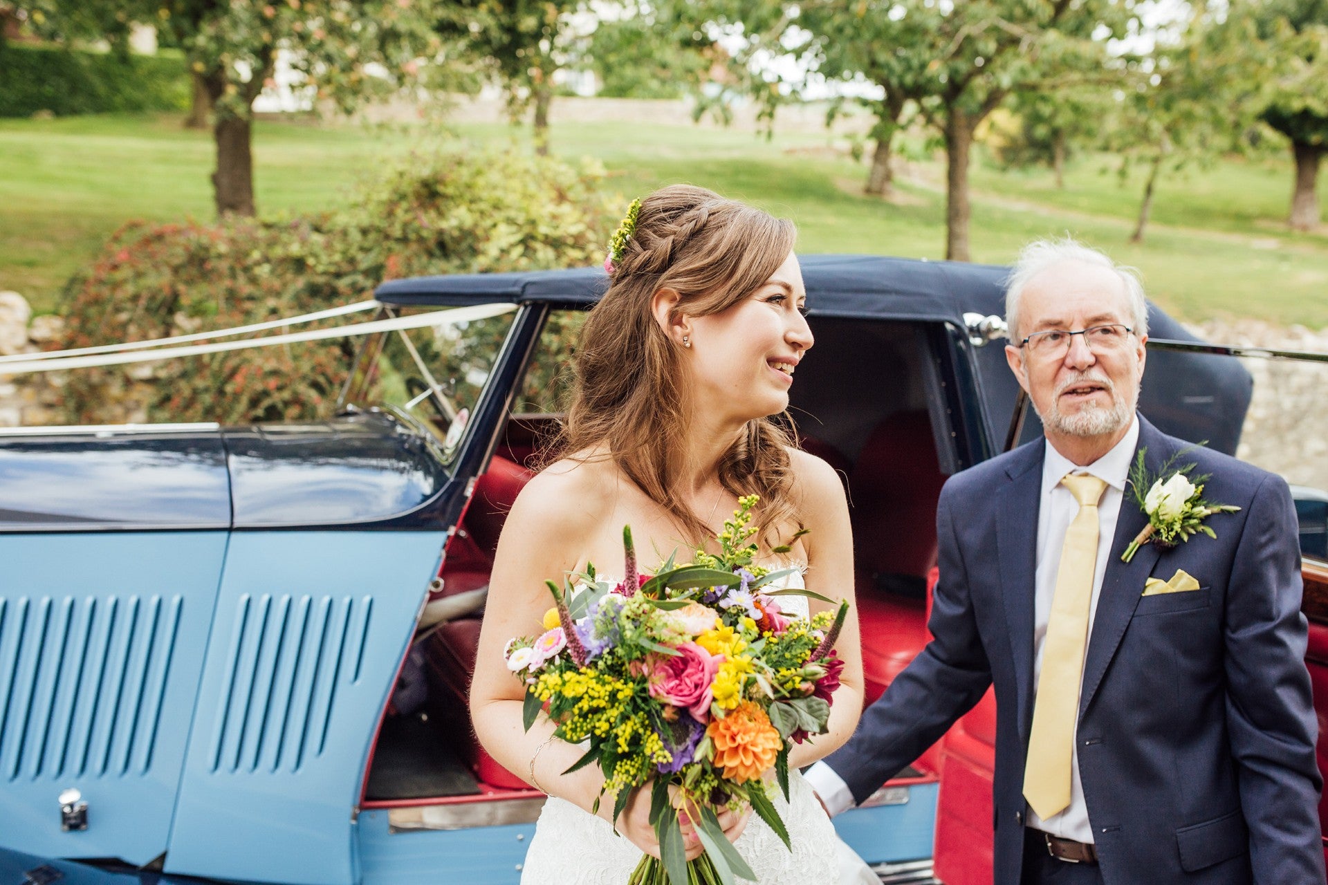 Bride holding a bouquet of flowers with her dad in front of a vintage car, in Bath, Somerset